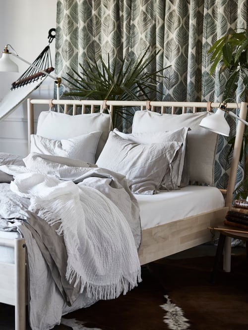 A BJÖRKSNÄS birch bed frame with a beige quilt cover, on top of a cowhide rug by a hammock in a bedroom with plants.