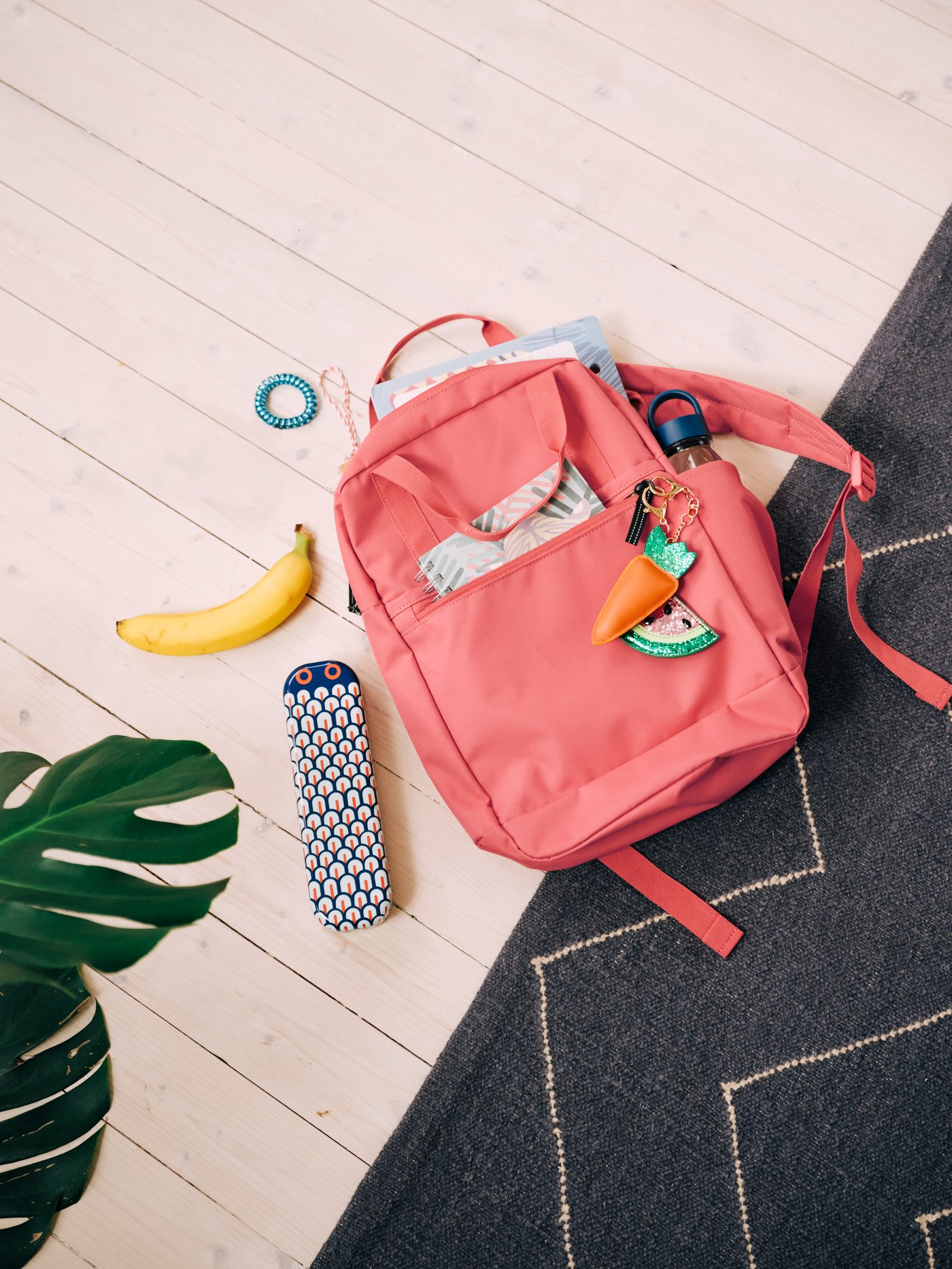 A pink-red STARTTID backpack on the floor, with a notebook and water bottle in the pockets and a pencil case beside it.