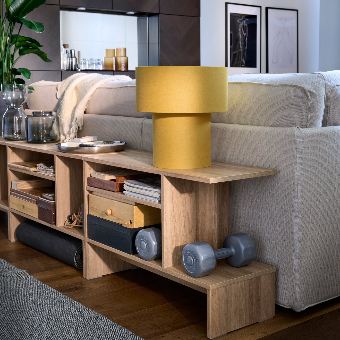 Mesas auxiliares HOLMERUD con contenido variado y una lámpara de mesa STOCKHOLM amarilla encima colocadas junto al respaldo de un sofá KIVIK.