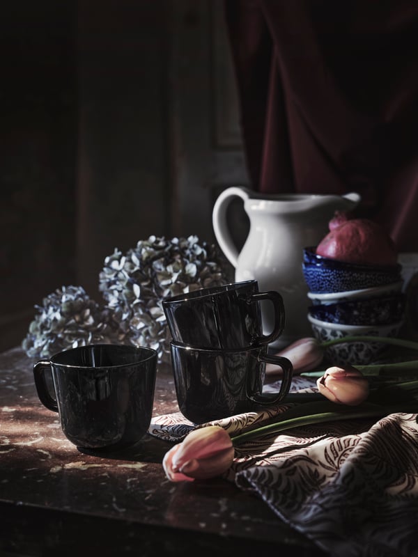 Three BACKIG mugs in glossy black glass placed on a countertop with other mugs and a jug, with some tulips lying nearby.
