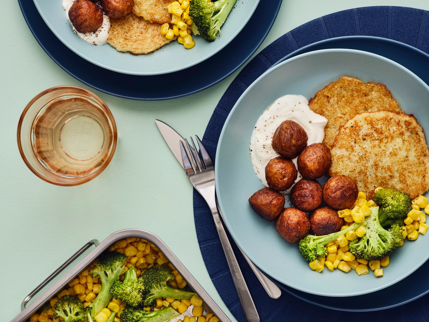 Two blue bowls filled with a meal of HUVUDROLL plant balls, potato pancakes and a mix of corn and broccoli are on a table.