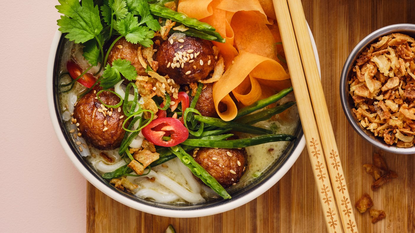 Three bowls containing dishes with Asian flavours are placed on a bamboo surface. Chopsticks are resting on one of the bowls.