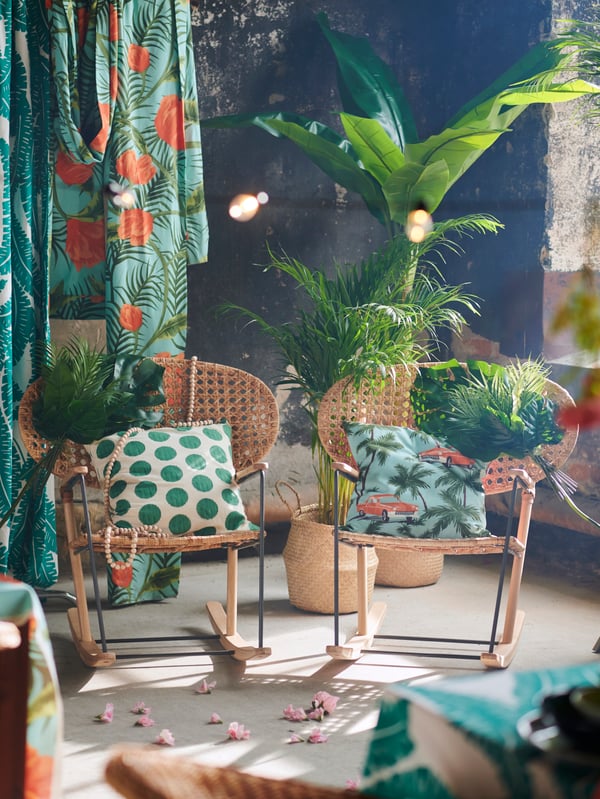 A pair of decorated GRÖNADAL rocking chairs side by side, surrounded by colorful textiles, tropical plants and flowers.