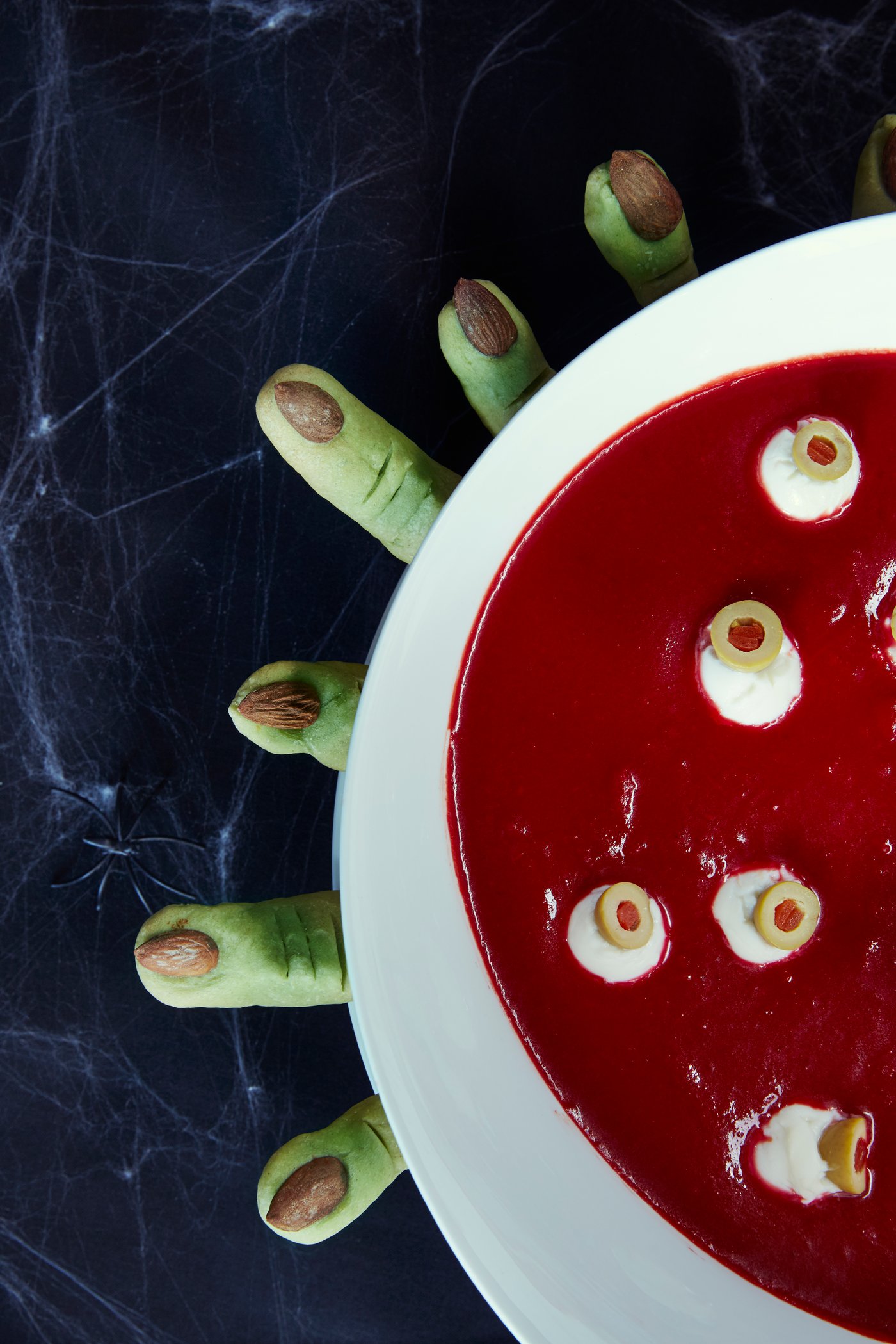 Et nærbilde av Halloween-mat som ligner blod med øyeepler som flyter i en hvit bolle med grønne småkaker som ligner fingrene rundt.