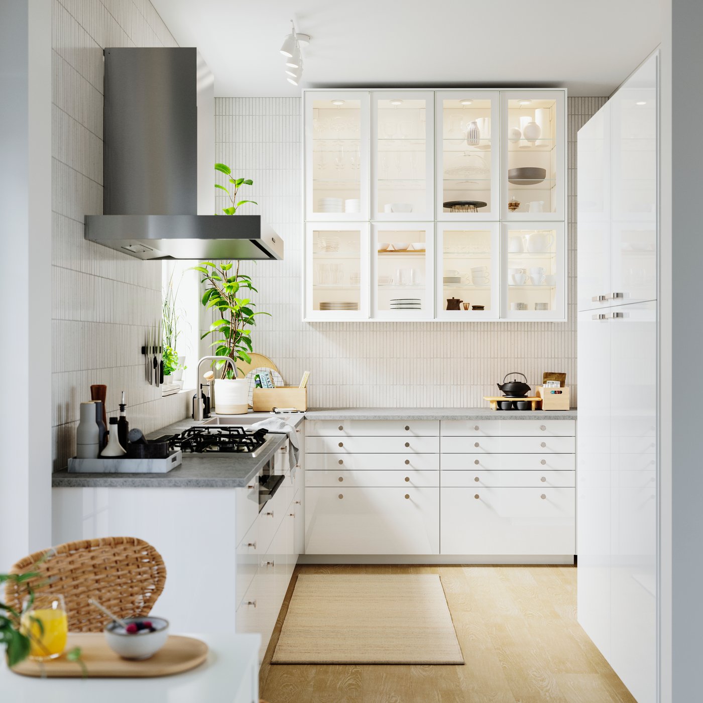 Moderna kuhinja s bijelim ormarićima i staklenim vratima, sivom radnom pločom i metalnom napom. Na pultu su biljke i kuhinjski pribor, a na podu svijetli tepih. Prirodno svjetlo osvjetljava prostor.