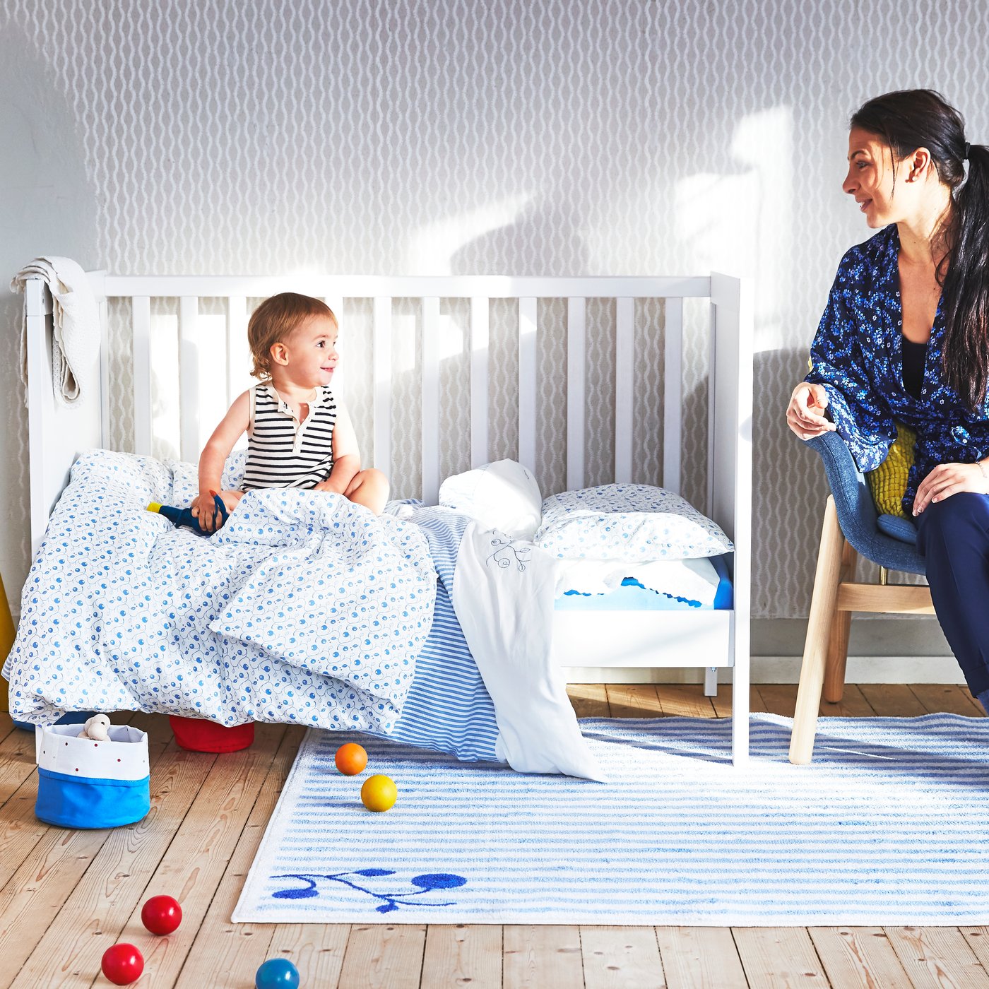 Mother sitting on a wooden chair and baby sitting in white open-sided cot with blue textiles both looking at each other.