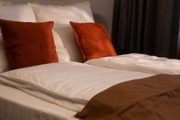 Zwei rotbraune DYTÅG Kissenbezüge in einem Bett mit weißer NATTJASMIN Bettwäsche und einer braunen DYTÅG Tagesdecke.