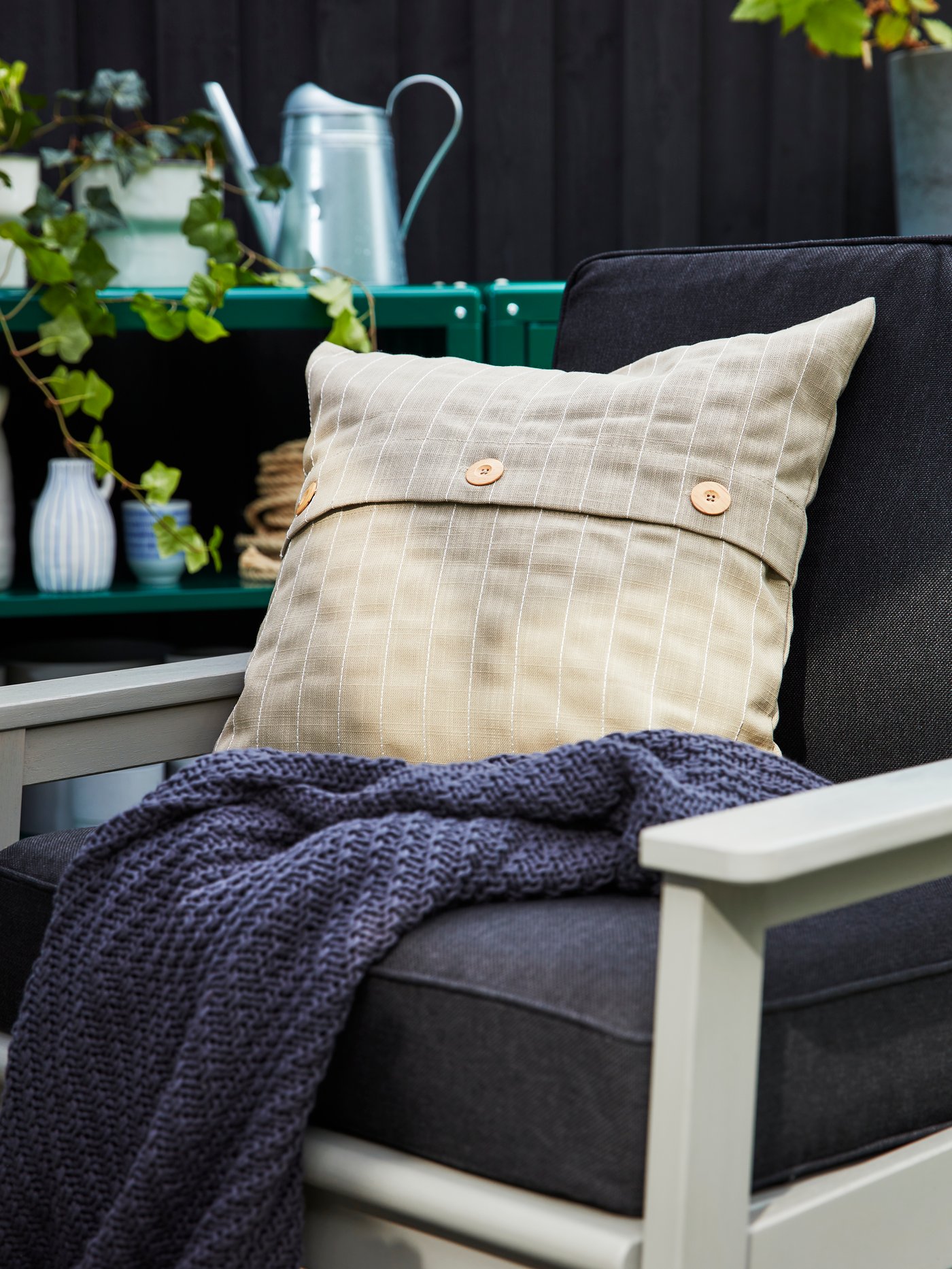 A grey outdoor armchair with back and seat cushions in dark grey, a beige outdoor cushion and a blue throw.