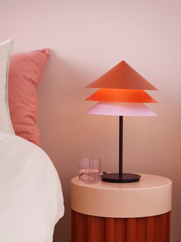 A TESAMMANS table lamp and glass are on top of a TESAMMANS side table beside a bed in a pink bedroom.