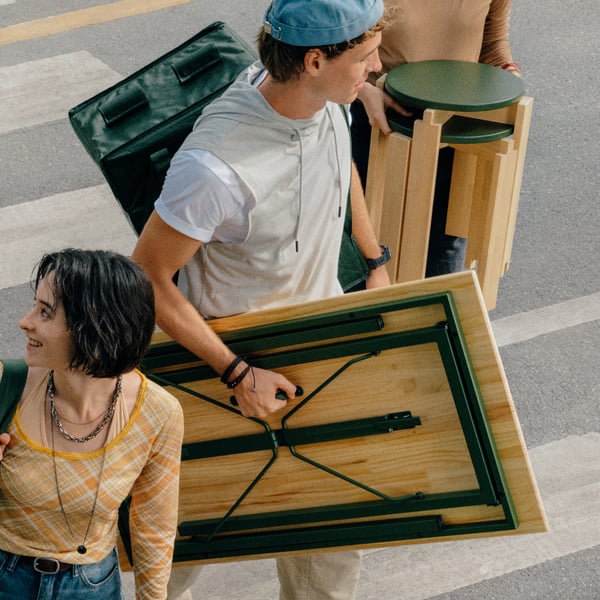 A person carries a folded KOMPISHÄNG desk by the handle across the street. KOMPISHÄNG hanging storage hangs on the shoulder.