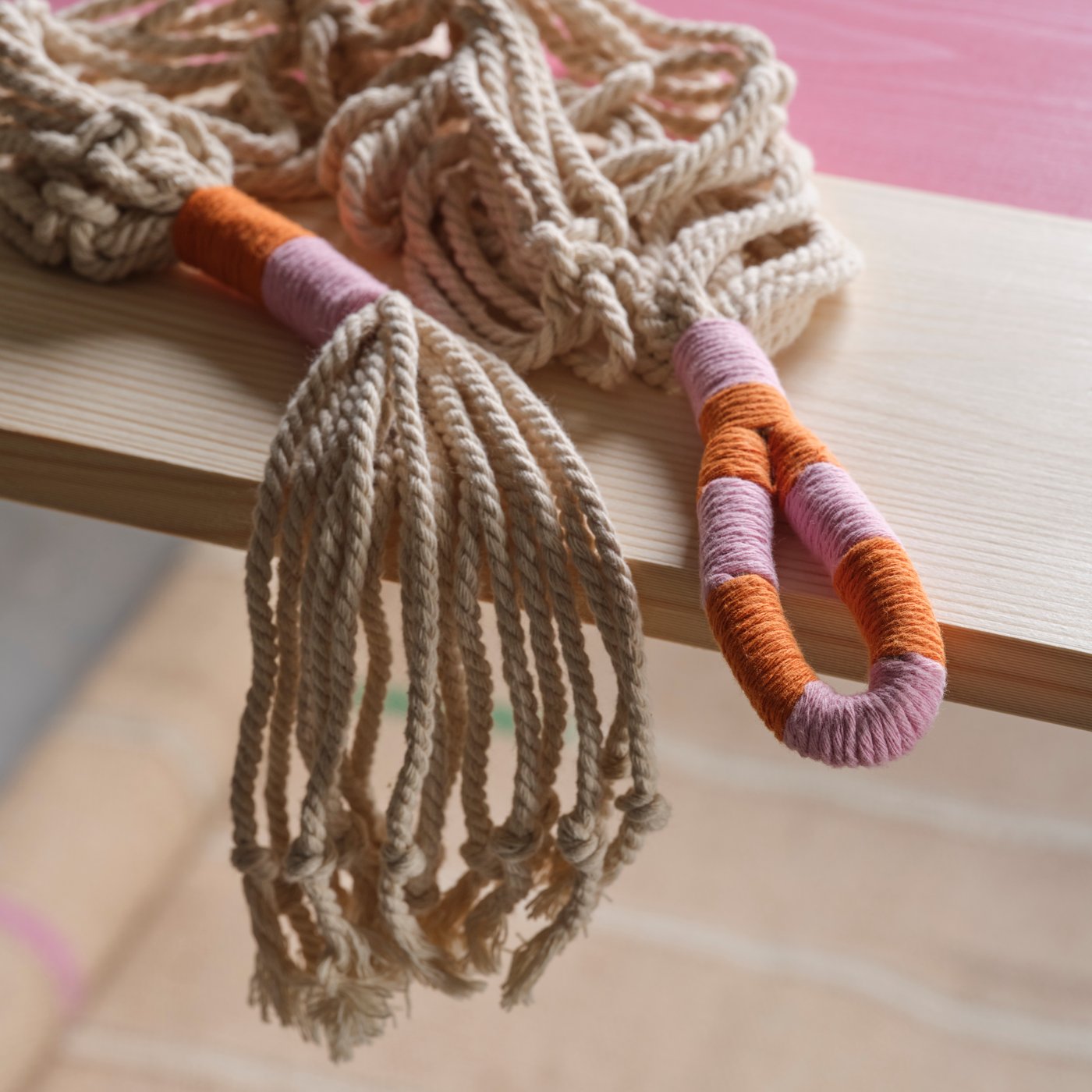 Un macetero colgante MÄVINN hecho a mano en macramé blanco con detalles en naranja y rosa sobre una tabla de madera.