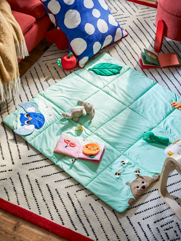 A SANDBI play mat and a cushion with a cover made from RAKLOSTA pre-cut fabric lie on an UNDERVISNING rug on the floor.
