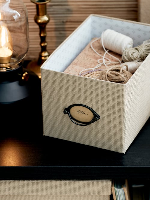 Close up on workspace table holding a rustic table lamp and a beige paper storage box with lid, containing sewing equipment. 