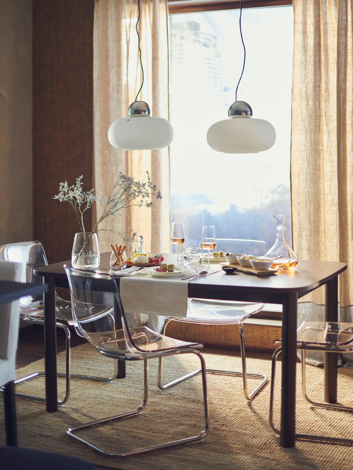 Coin repas avec une table en bois dressée, des chaises transparentes, deux suspensions lumineuses et de la vaisselle, devant une grande fenêtre avec des rideaux légers.