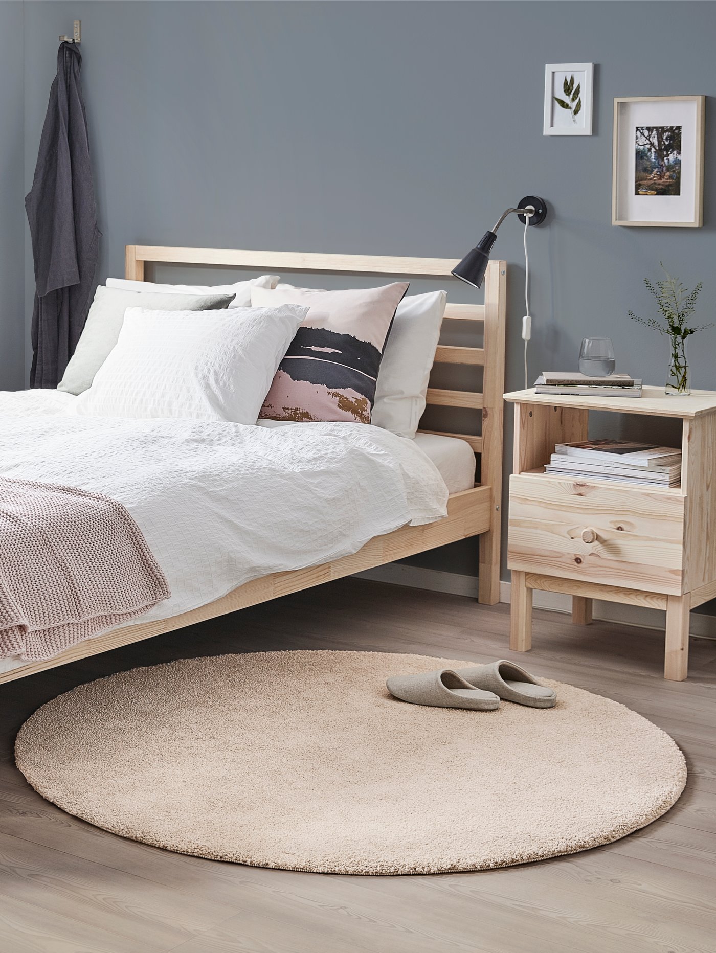Grått sovrum med en säng i furu bäddad med vita sängkläder. Intill står ett nattduksbord i furu och på golvet ligger en rund STOENSE matta i beige.