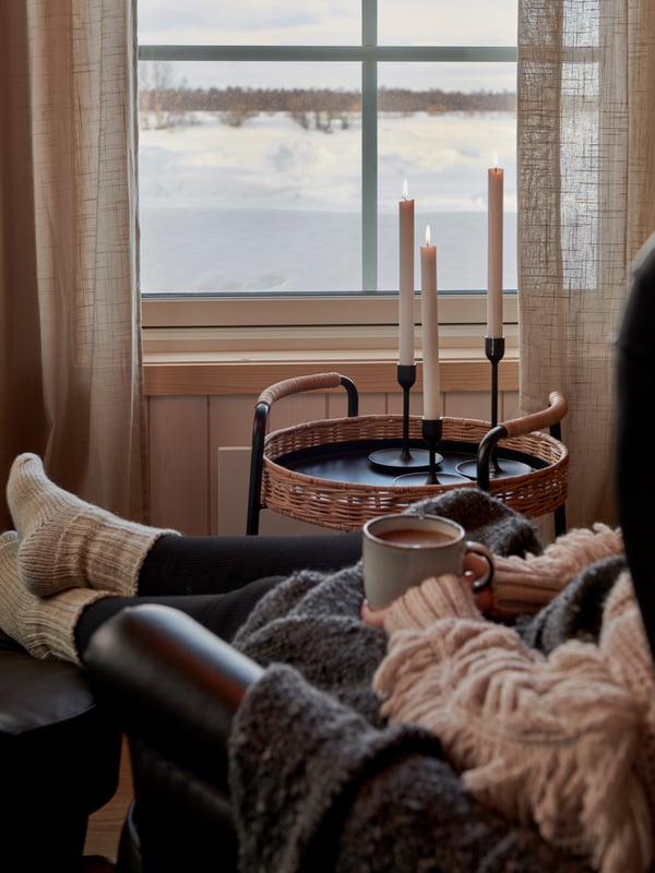 Person med raggsockor som sitter vid ett fönster med snö utanför, håller en mugg. På ett litet bord bredvid står tända ljus.