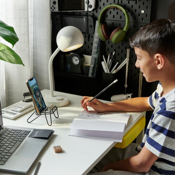 A young boy sits at a white ENKROKEN desk, with a laptop, notebook, and a white KORVSNÖRE LED lamp on top.