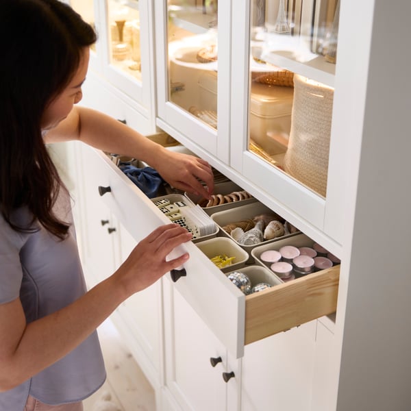 A person has pulled out the drawer in an IDANÄS high cabinet with glass doors and 1 drawer, reaching for items stored inside.