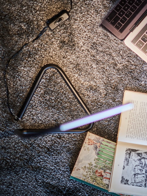 A HÅRTE LED work lamp in black/silver-colour with a triangular base and thin body illuminates a laptop and books on a floor.
