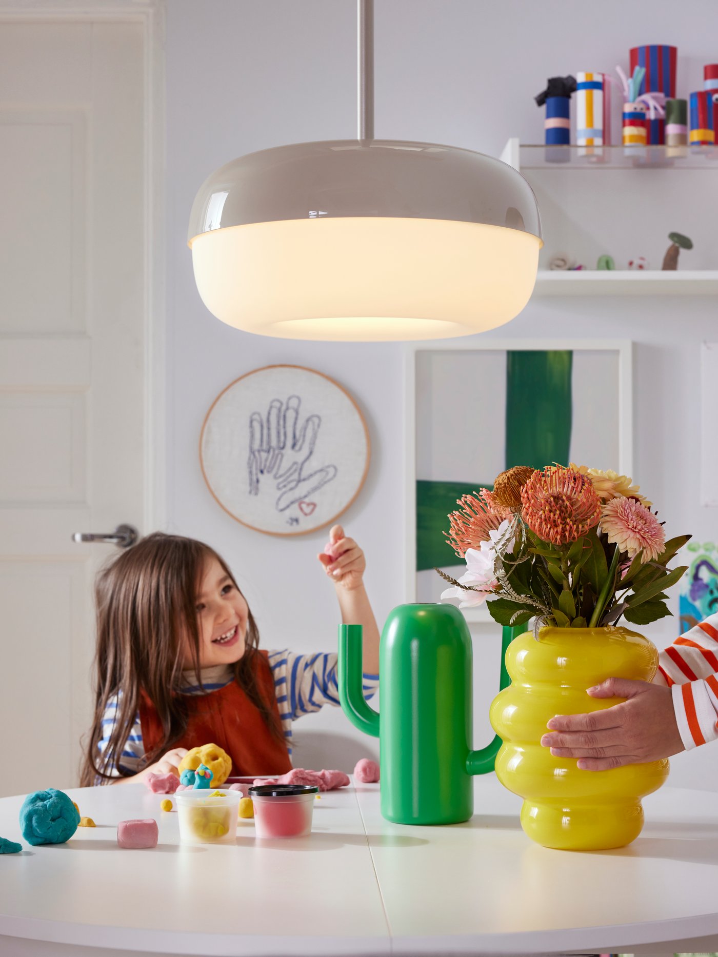 A beige BLÅSVERK pendant lamp hangs over a table with a yellow KÄLLARHALS vase and an ÄRTBUSKE vase/watering can on it.