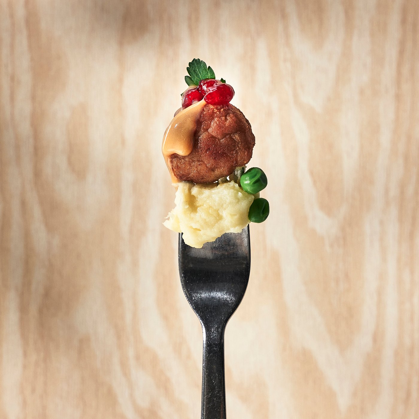 A fork holding mashed potatoes, green peas and a meatball topped with lingonberry sauce and parsley against a light wooden background.