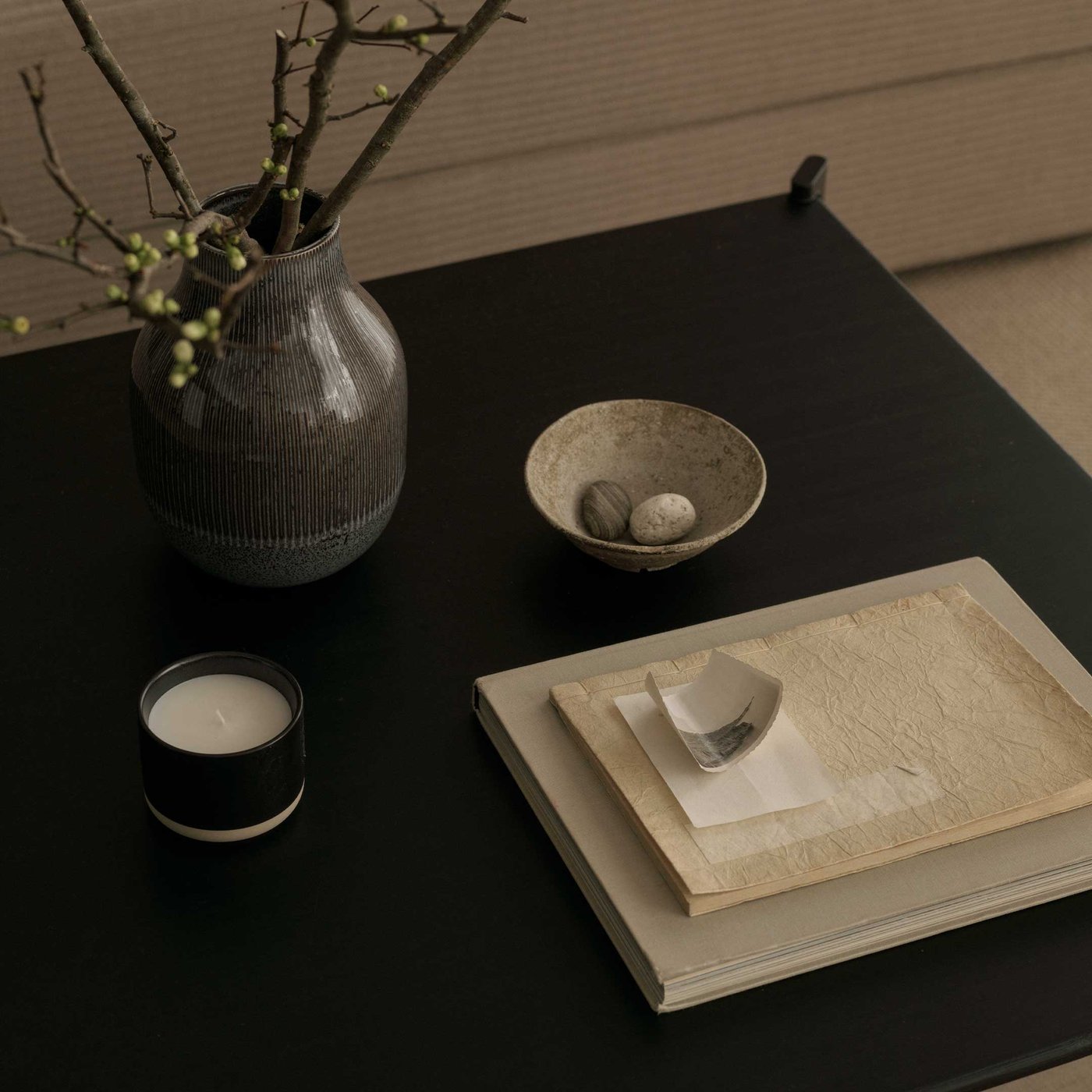 A beautifully styled dark surface, perfect for adding a touch of natural elegance and calm to your home. A ribbed, dark grey vase holds delicate branches with fresh green buds, bringing a hint of the outdoors in. Nearby, a small, textured bowl cradles two smooth pebbles, adding a touch of natural serenity. A cosy candle in a black holder with a white base promises a warm glow for a relaxing evening. A stack of books and papers, topped with a small, curved decorative object, suggests a quiet moment for reflection. This arrangement creates an inviting and peaceful atmosphere, showcasing how thoughtful details can transform a space.