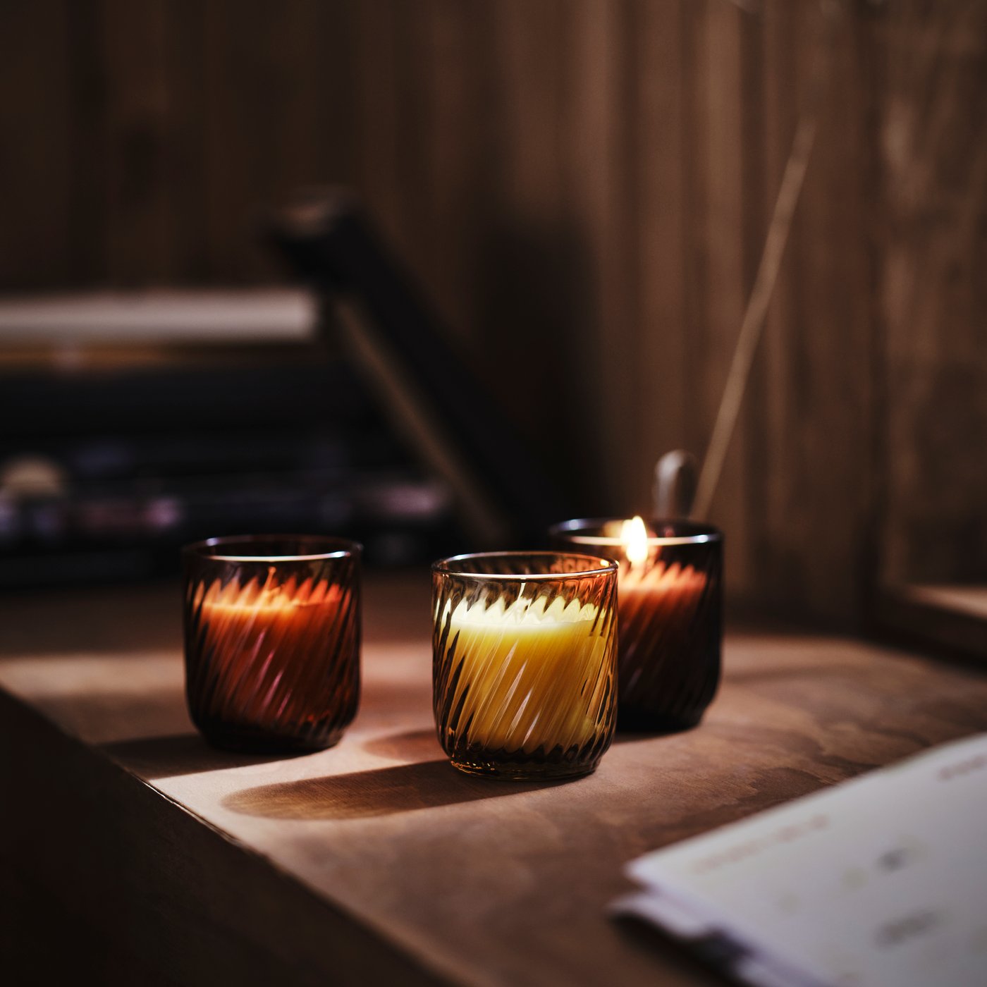 A close-up of three TÖRNFÅGEL glass candle holders in brown and green in a dimly lit room on a wooden surface.