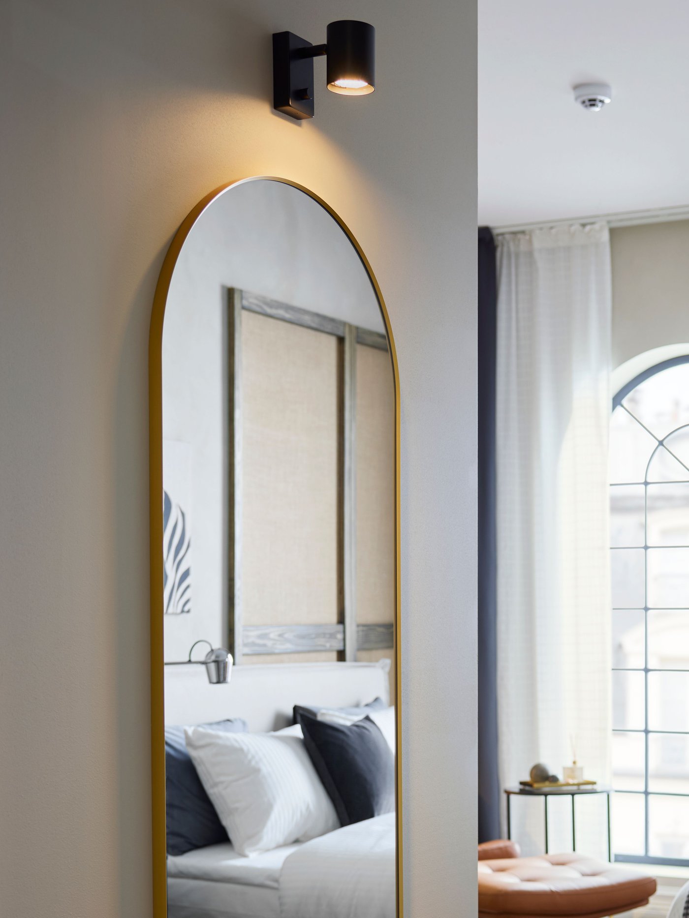 Under a lit wall lamp is a LINDBYN mirror with a gold-coloured frame that is rounded on top, partly reflecting a hotel bed.