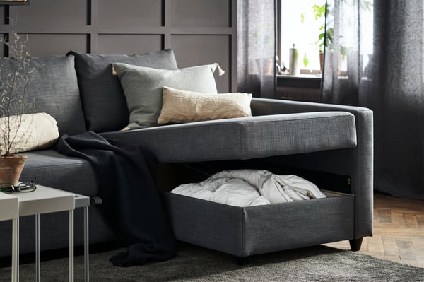 Divano modulare grigio scuro con modulo reclinabile e vano portaoggetti estraibile contenente biancheria. Appoggiati sul divano ci sono dei cuscini decorativi, una coperta scura e si trova in un soggiorno con tende grigie e piante