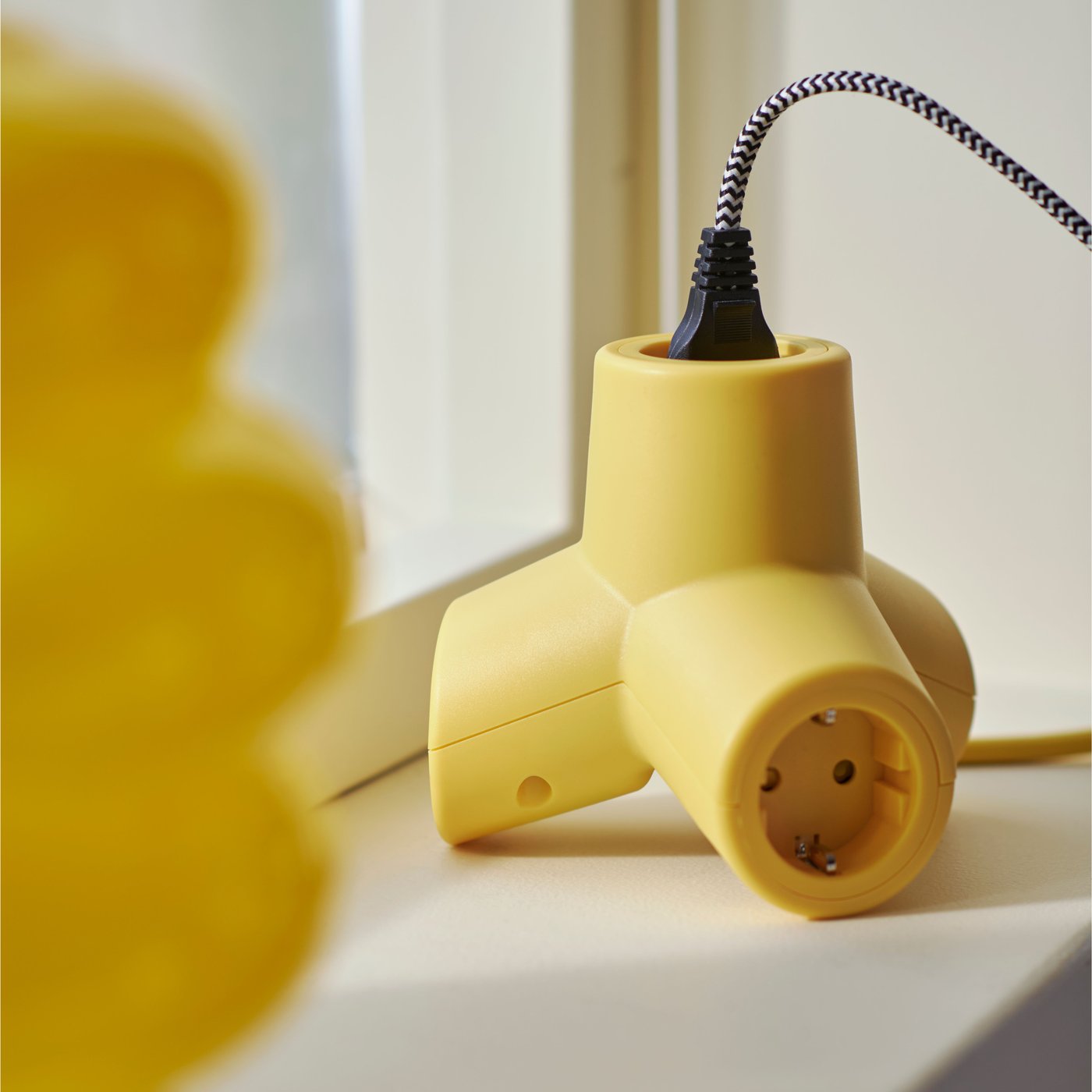 Žlutý prodlužovací kabel HORNFELS se čtyřmi zásuvkami leží na okenním parapetu vedle žluté vázy a jeden z jeho kabelů je zapojený do zásuvky.