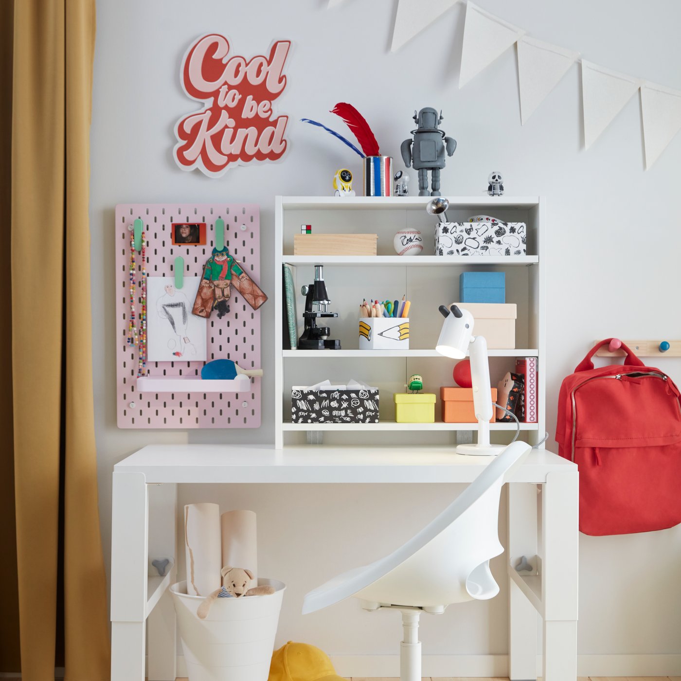 Skrivbord med hylla, böcker, robotfigur, röd ryggsäck och anslagstavla med skylten 'Cool to be Kind' i ett ljust rum med trägolv.