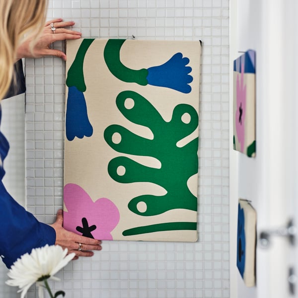 A person adjusts some multicoloured FJÄLLDRABA pre-cut fabric which is being stapled onto a rectangular board.