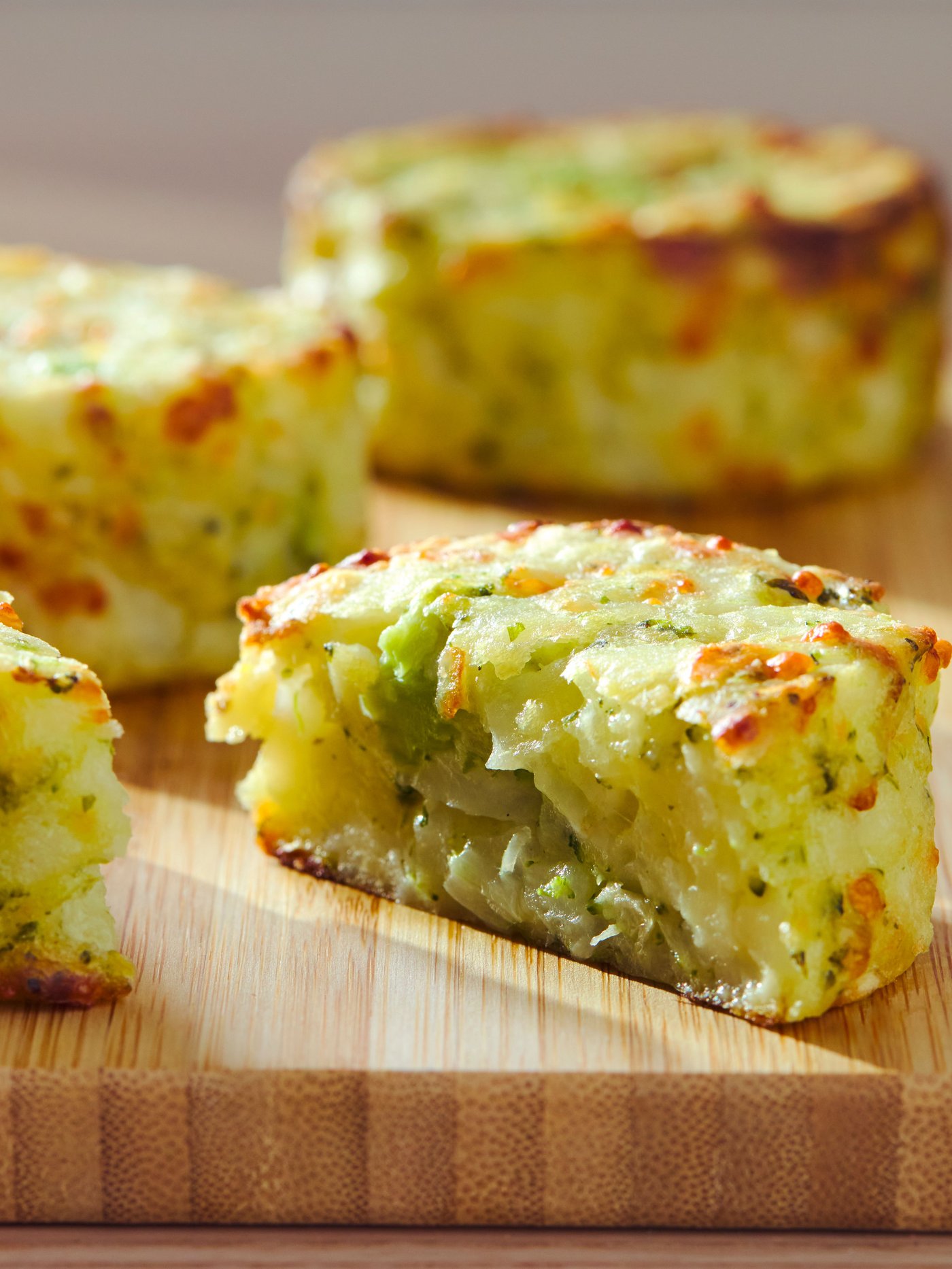 Krupan kadar tri zlatno-braon kolača od krompira na drvenoj dasci, s vidljivim zelenim biljem i crvenim mrljama. U prvom planu je kolač presečen na pola, čime je otkrivena meka unutrašnjost iz koje izlazi para