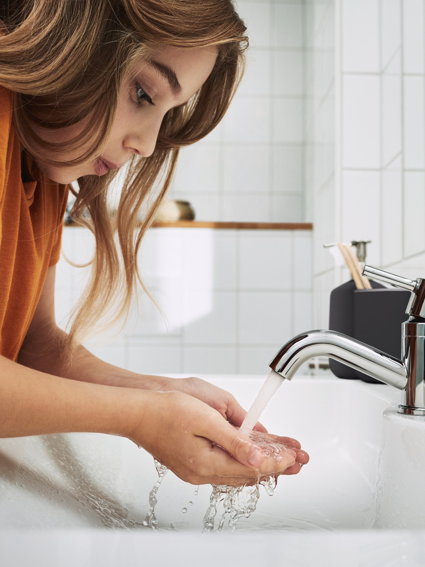 A young child with long brown hair standing in a white tiled bathroom rinsing their hands in the wash basin.