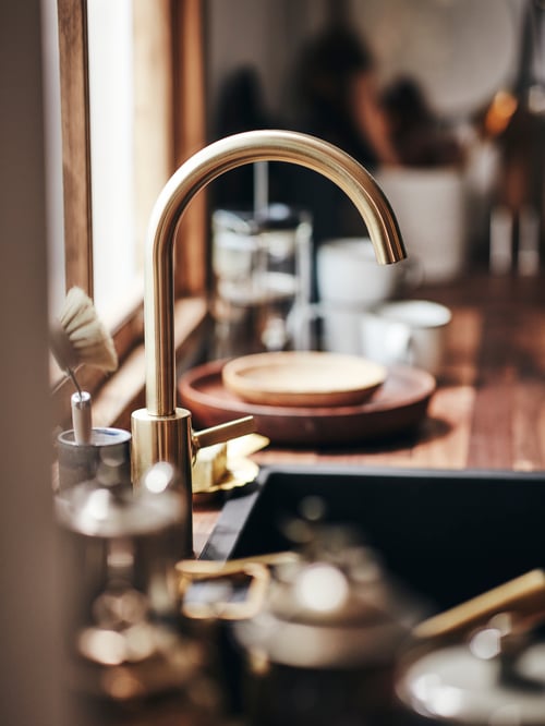 A close-up of a DELSJÖN kitchen tap above a sink with dishes and cups in the background.