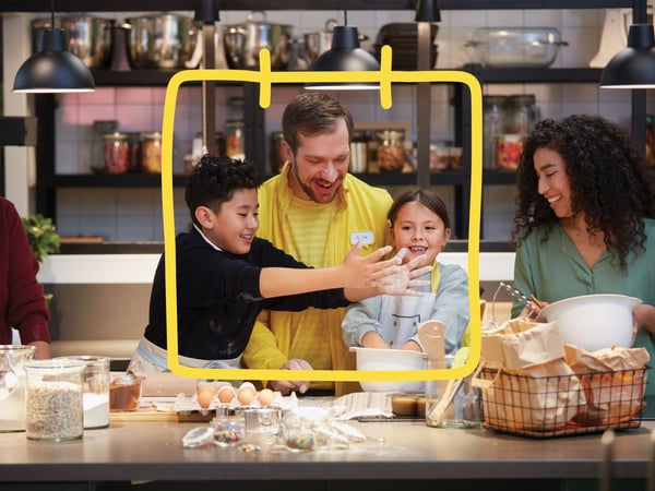 IKEA co-worker in a yellow sweater baking together with two children and an adult around a kitchen counter with baking ingredients in an IKEA showroom kitchen.