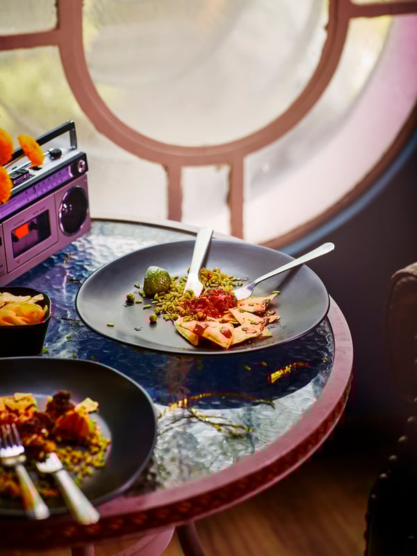El borde de una mesa redonda con superficie de vidrio. Hay platos con restos de comida y detrás una radio de los ochenta.