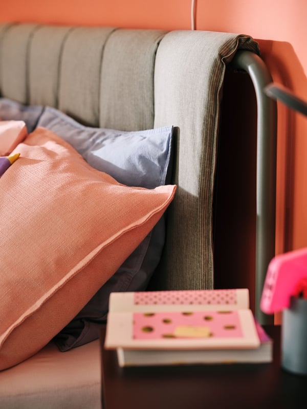 A pillow in a grey blue ÄNGSLILJA pillowcase and a cushion in a pink LAGERPOPPEL cover lean against an upholstered headboard.