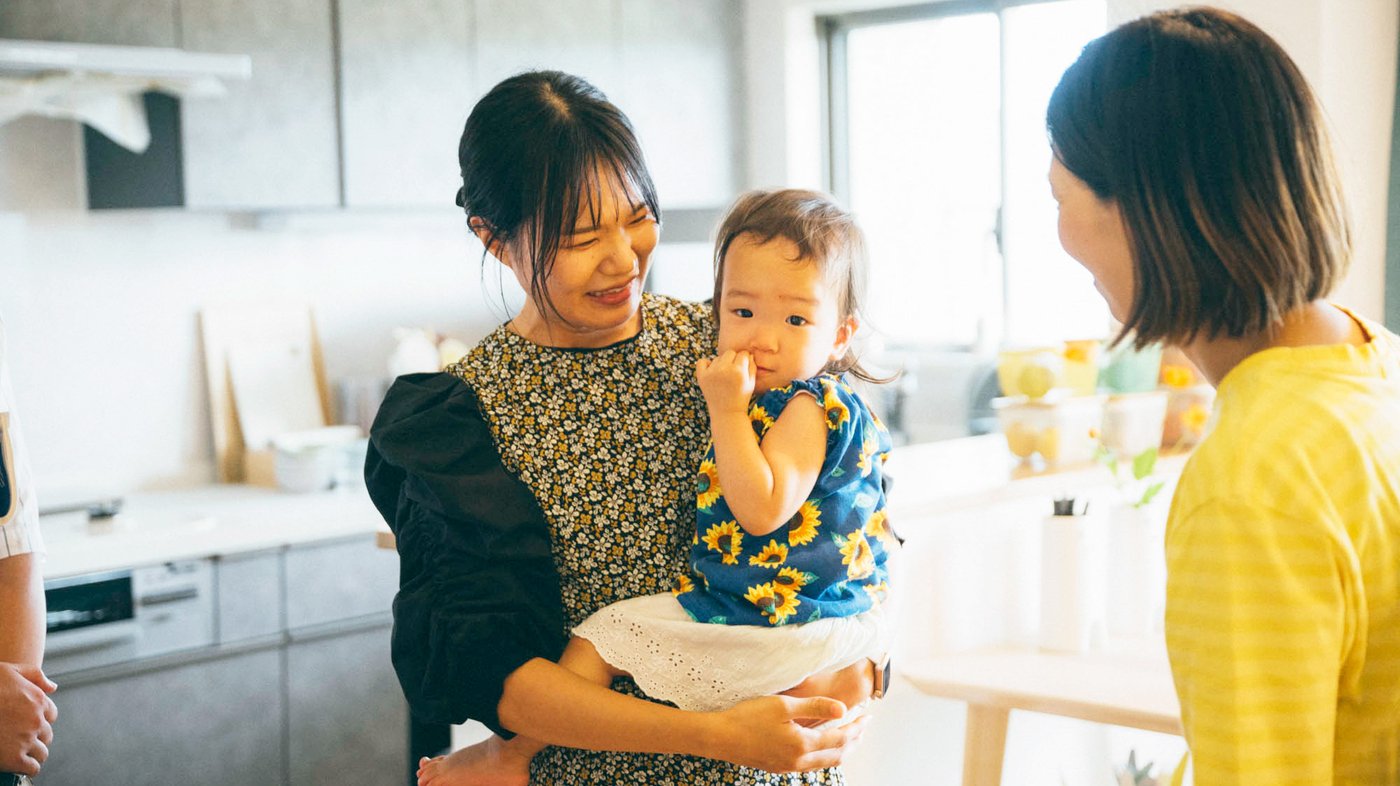 現代的なキッチンで、子どもを抱いた人物と、もう一人の人物が交流している様子。キッチンのカウンターにはさまざまなアイテムが置かれ、奥に窓があります