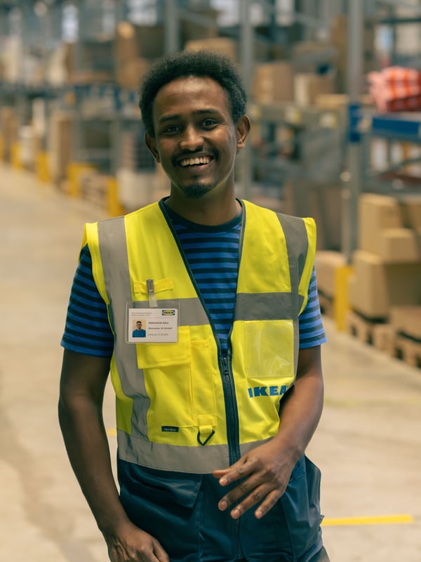 Abdi wearing a reflective safety vest and shirt walks through a large warehouse filled with organized shelves and cardboard boxes