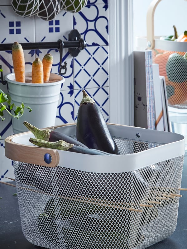 A white metal bin on a countertop, holding an eggplant and three carrots.