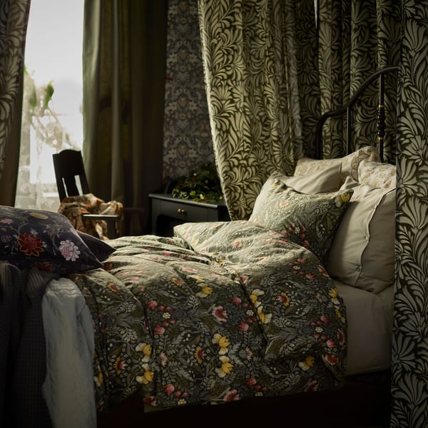 Dark green/multicolour KÄRRDUNÖRT bed linen on a bed that’s surrounded by patterned curtains, near a window.