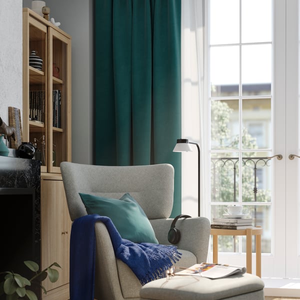 A living room featuring a sunlit beige-gray OSKARSHAMN wing chair and storage footstool, placed by French windows dressed with teal-blue curtains layered over a sheer white one.