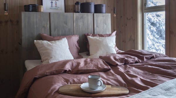 En dobbelseng redd opp med puter og dyner kledd i dus rosa sengesett, beige putetrekk og en gavl bak sengen laget av to IVAR skap som er beiset.