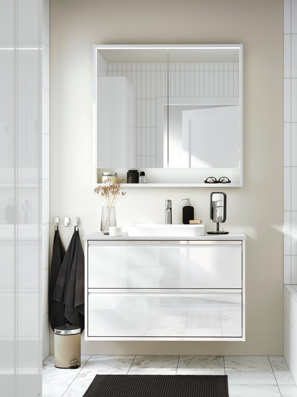 An ÄNGSJÖN vanity sink with drawers in high-gloss white has a white mirror cabinet above it and two towels on hooks next to it.