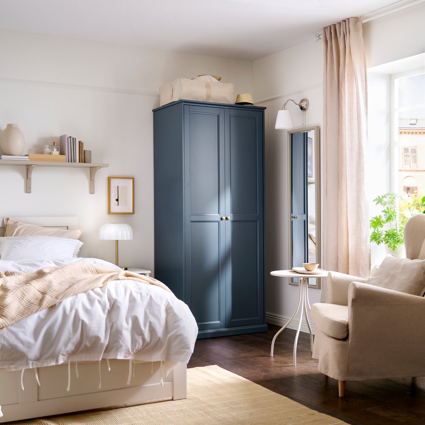 Un dormitorio luminoso con un armario FÅGELFJÄLLET azul marino junto a una cama con ropa de cama en tonos blanco y beige.