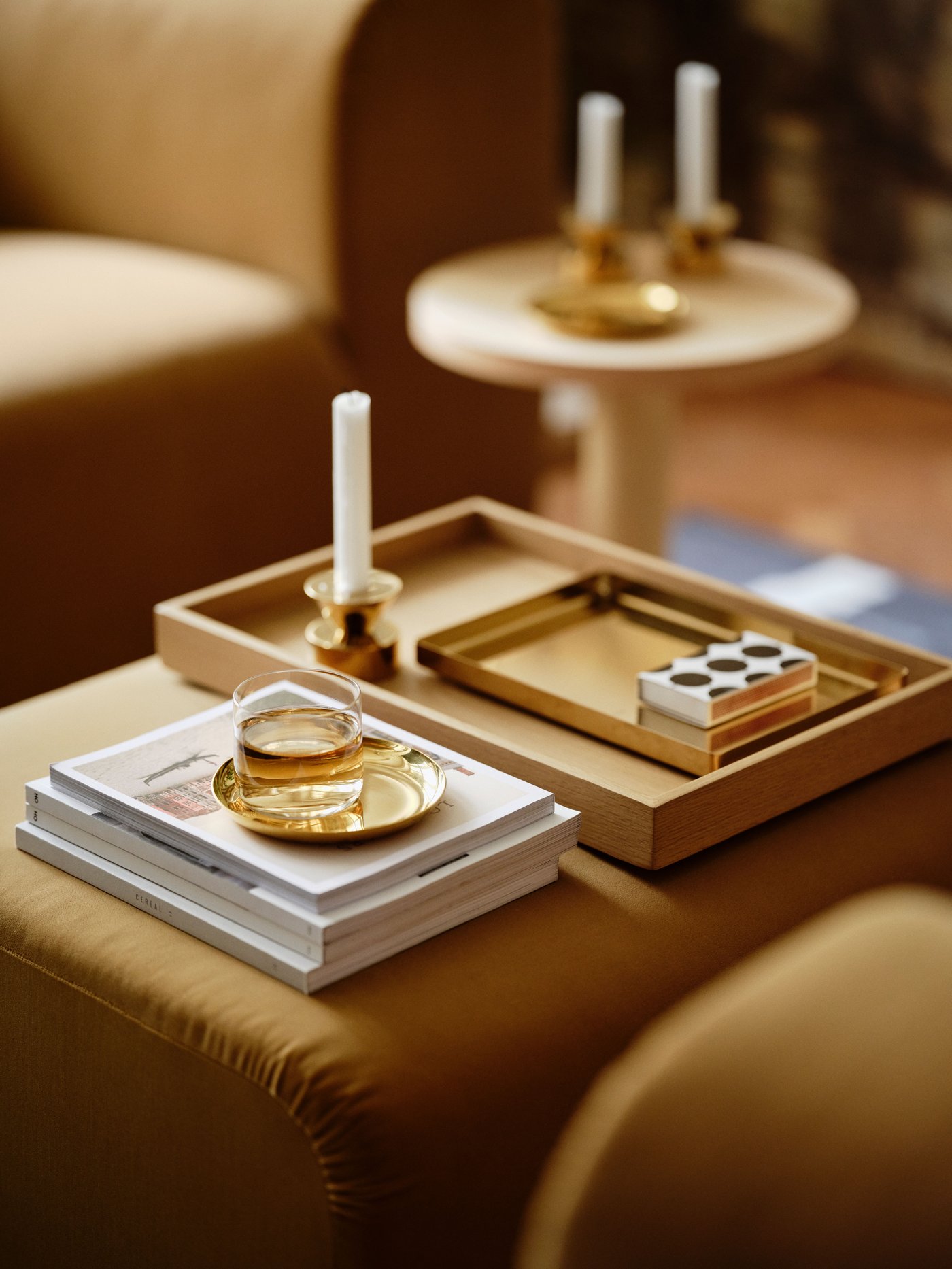 Un vassoio STOCKHOLM 2025 con all'interno un vassoio di dimensioni più piccole in ottone, un portacandele e altri oggetti decorativi, posti sopra un pouf.
