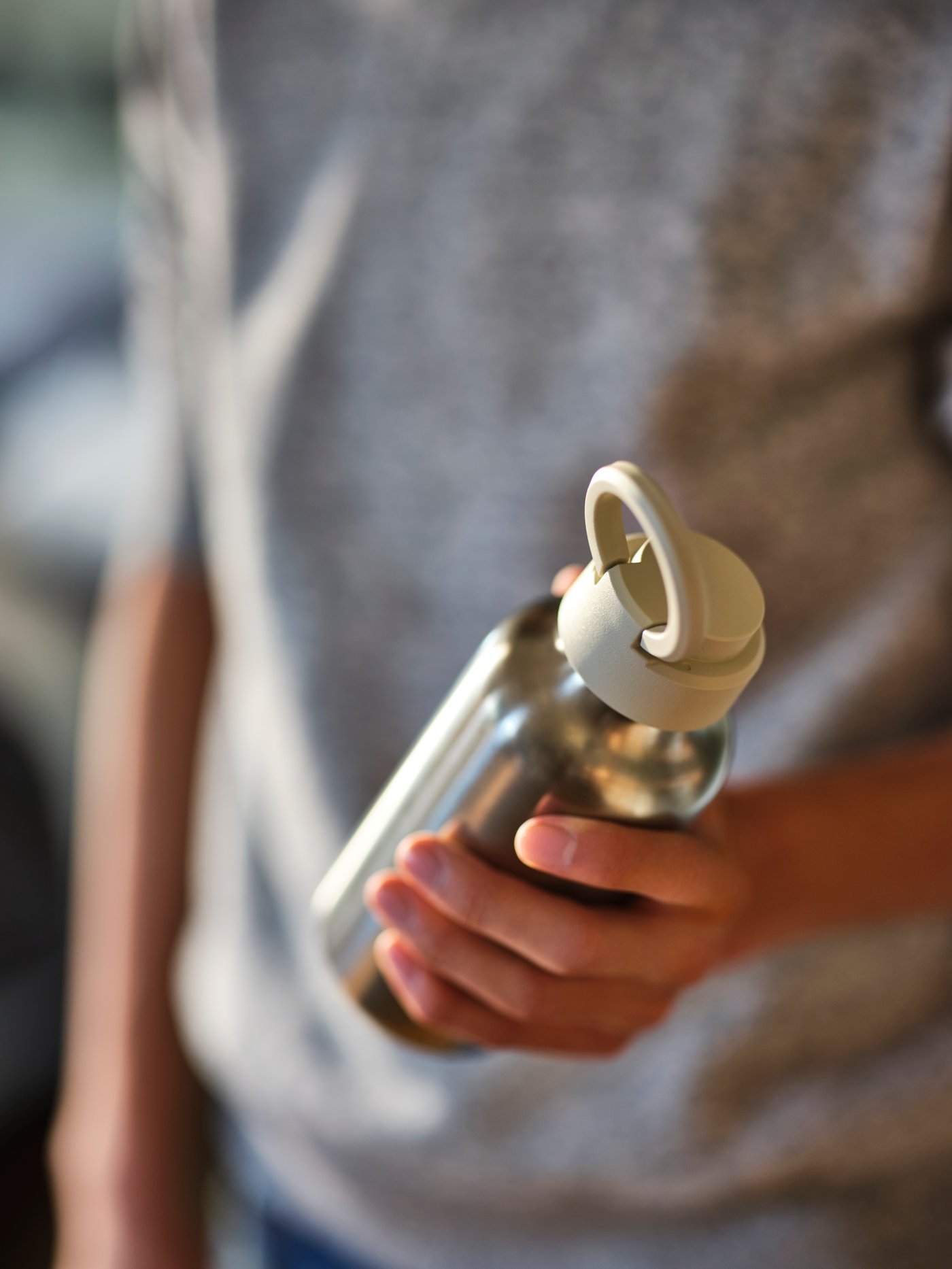 Osoba w szarej bluzie trzymająca w ręku stalową butelkę na wodę ENKELSPÅRIG z beżową zaketką.