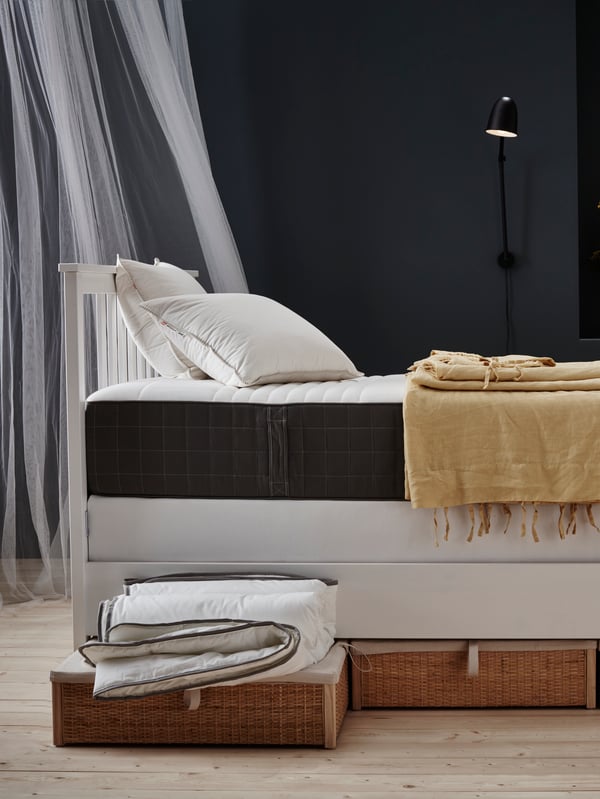 A white divan bed with a yellow throw and a rattan storage box under the bed. One rattan storage box has a duvet resting on it.