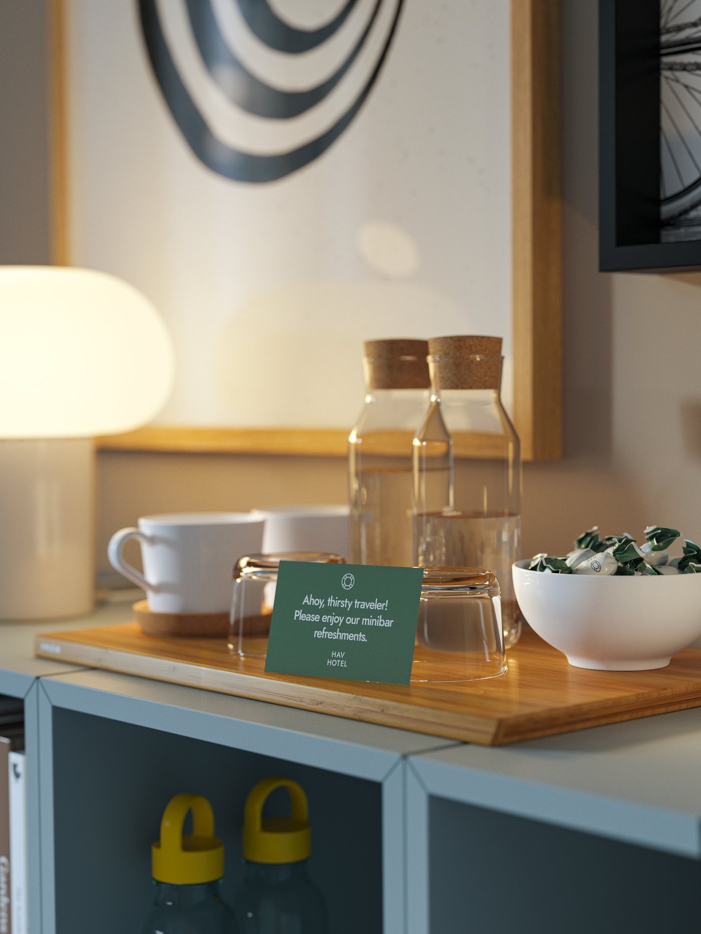 Dryckeshörna på en byrå med kopp, vattenflaskor, snacks och en informationsskylt, upplyst av en bordslampa och omgiven av inramad väggkonst.